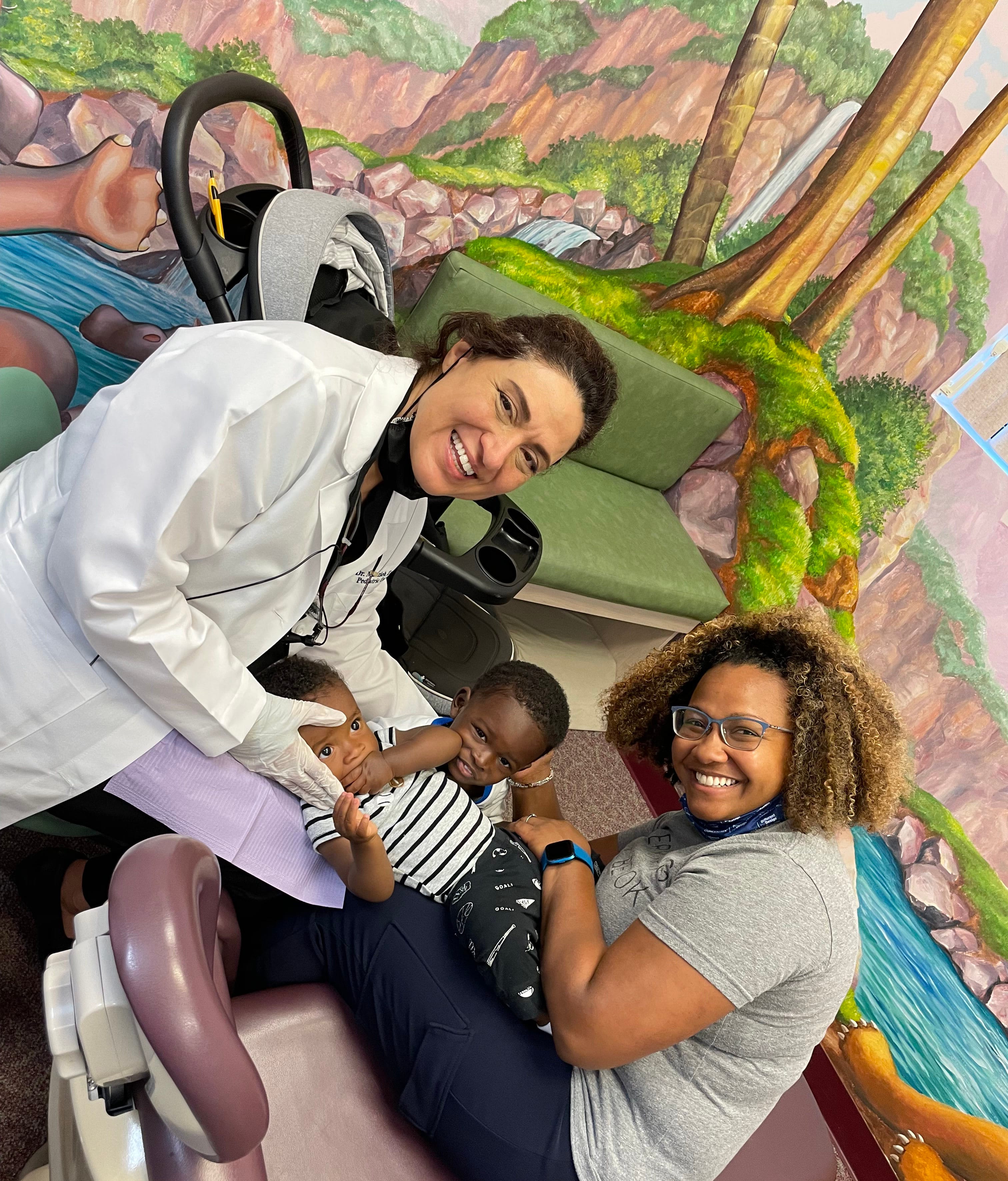 Dr Zahedi smiling along side a young african-american mother with her two young children in a pediatric dentistry office